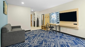A hotel room with a grey sofa, a TV mounted on the wall, a patterned carpet, and a wooden desk with a chair. - Baymont by Wyndham Grovetown Augusta (Grovetown)