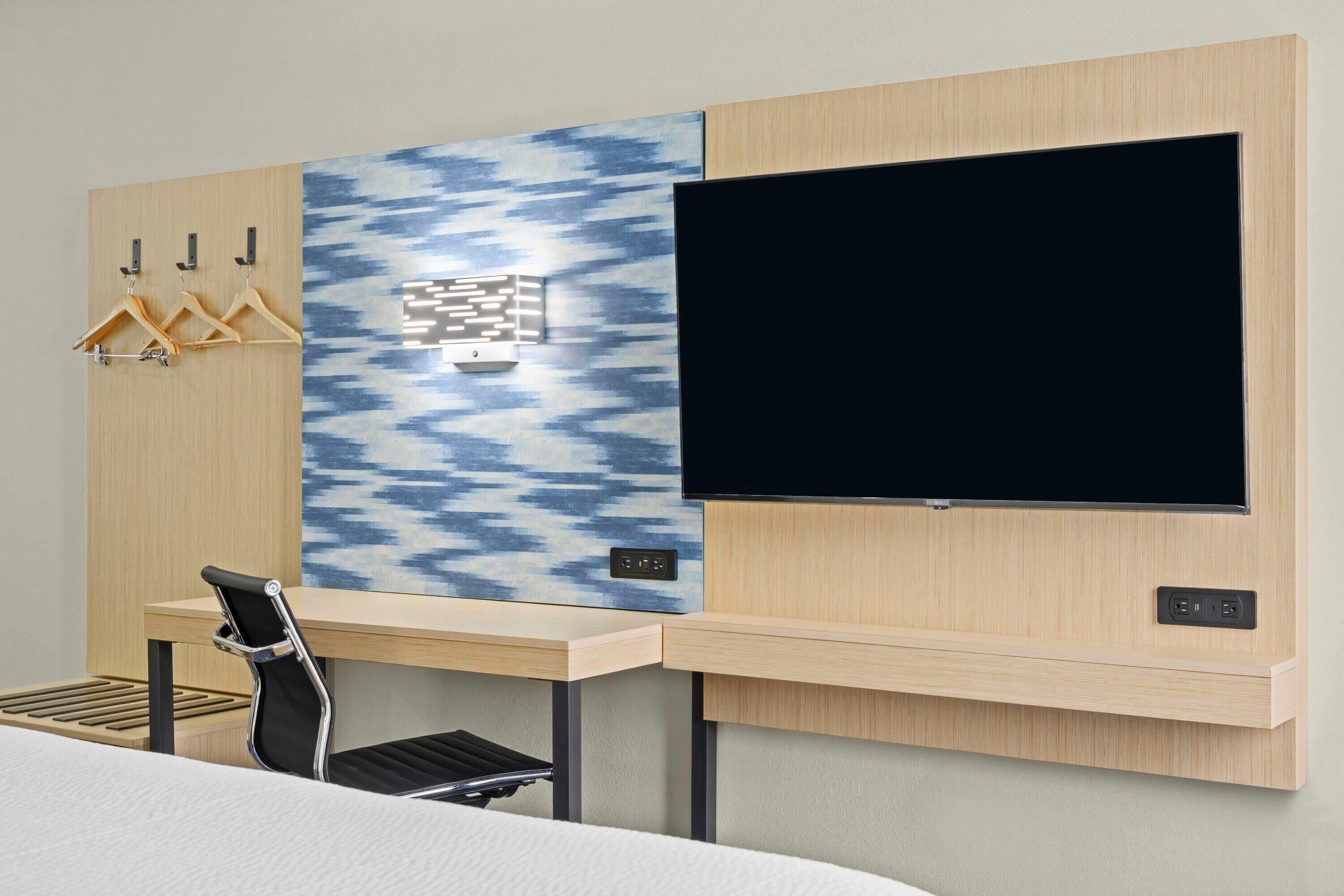 A hotel room with a bed, a desk with a chair, a TV mounted on the wall, and a coat rack with hangers.