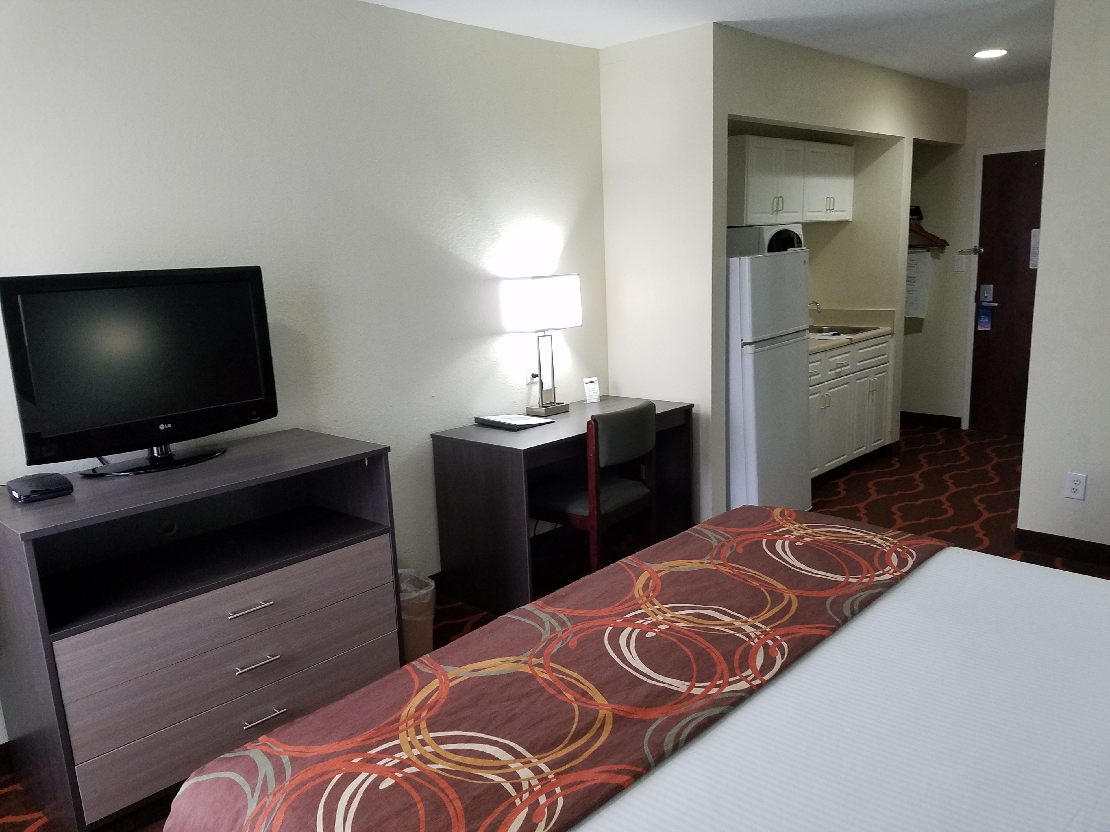 A hotel room with a bed, a TV, a desk with a lamp, and a kitchenette.