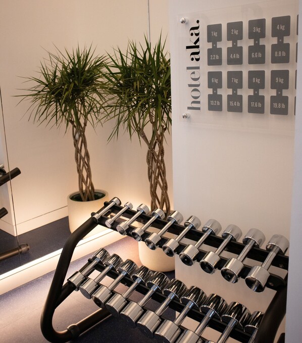 A hotel room with a wall-mounted scale displaying weight measurements in kilograms and pounds, a set of dumbbells, and two potted plants. - Hotel AKA Back Bay (Boston)