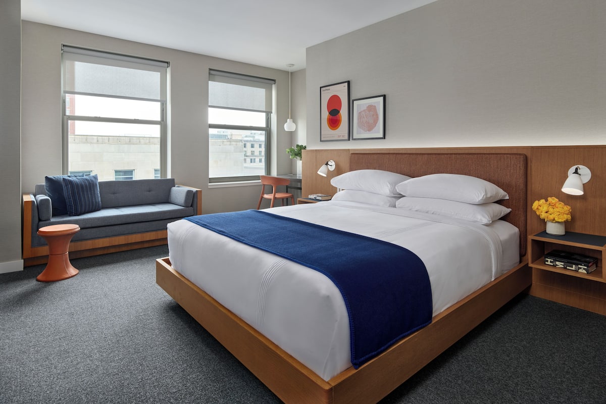 A modern hotel room with a large bed, a sofa, a desk, and a view of the city.