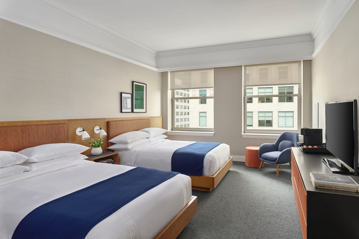 A hotel room with two beds, a desk, a television, and a window with a city view.