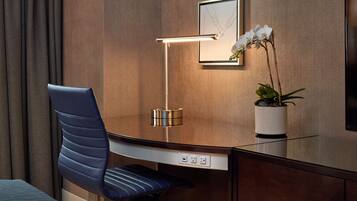 Una habitación de hotel moderna con un escritorio elegante, una silla azul, una lámpara de pie, una planta en maceta y una obra de arte enmarcada en la pared.