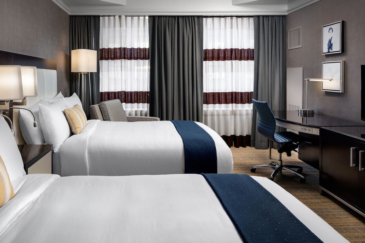 A hotel room with two beds, a desk, a chair, a TV, and a large window with curtains.