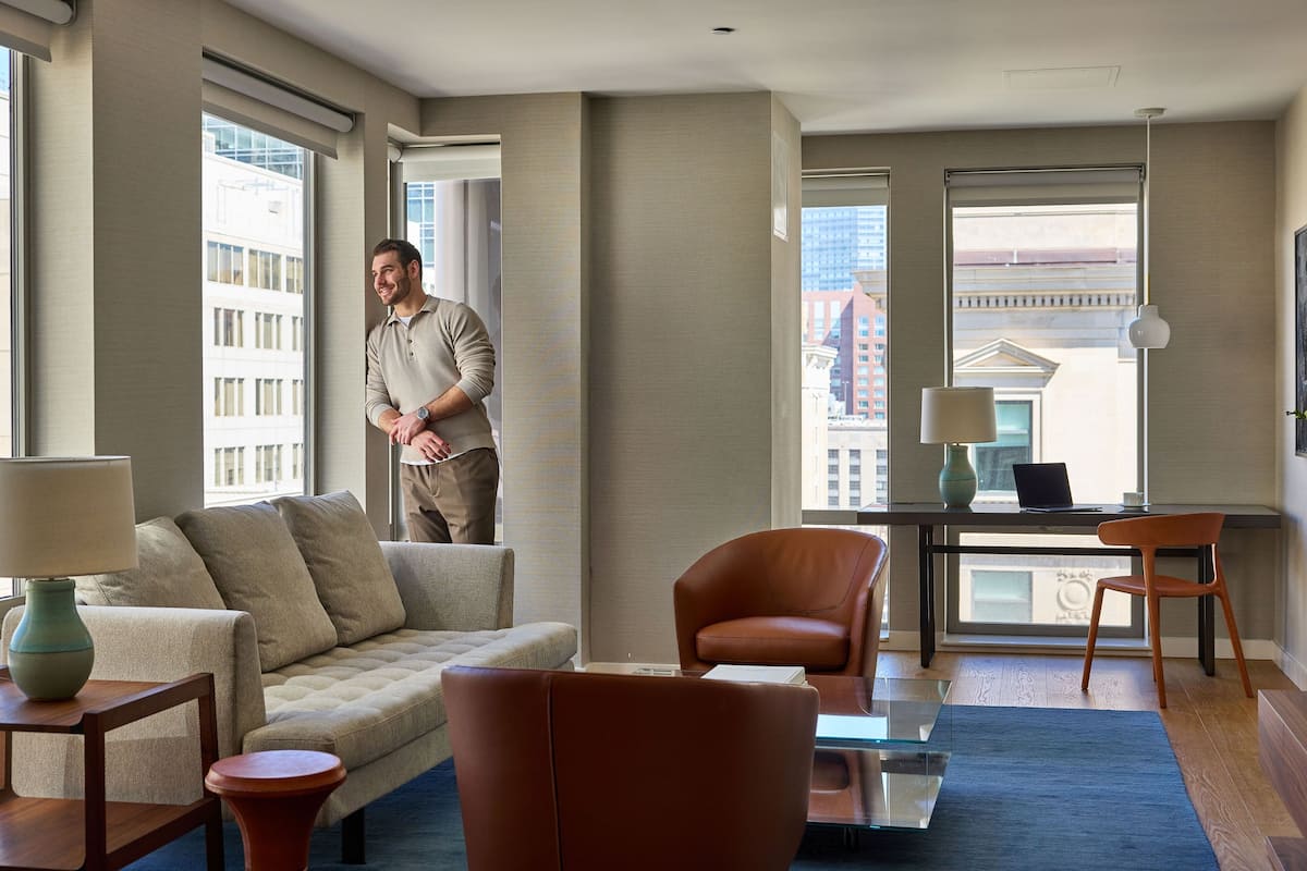 A modern hotel room with a large window, a sofa, a chair, a desk with a laptop, and a lamp.