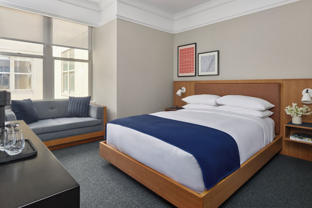 A hotel room with a bed, a sofa, a desk with glasses, and a nightstand with a lamp.