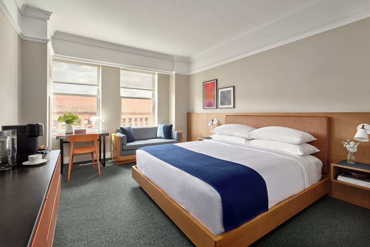 A hotel room with a large bed, a desk with a coffee maker, a chair, and a window with a view of buildings.