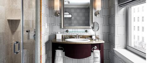 A modern bathroom with a glass shower enclosure, a vanity with a marble countertop, a large mirror, and a window with a view of buildings.