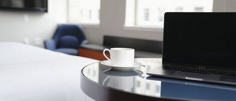 A laptop on a glass table with a white cup beside it, a bed, and a chair in a hotel room.