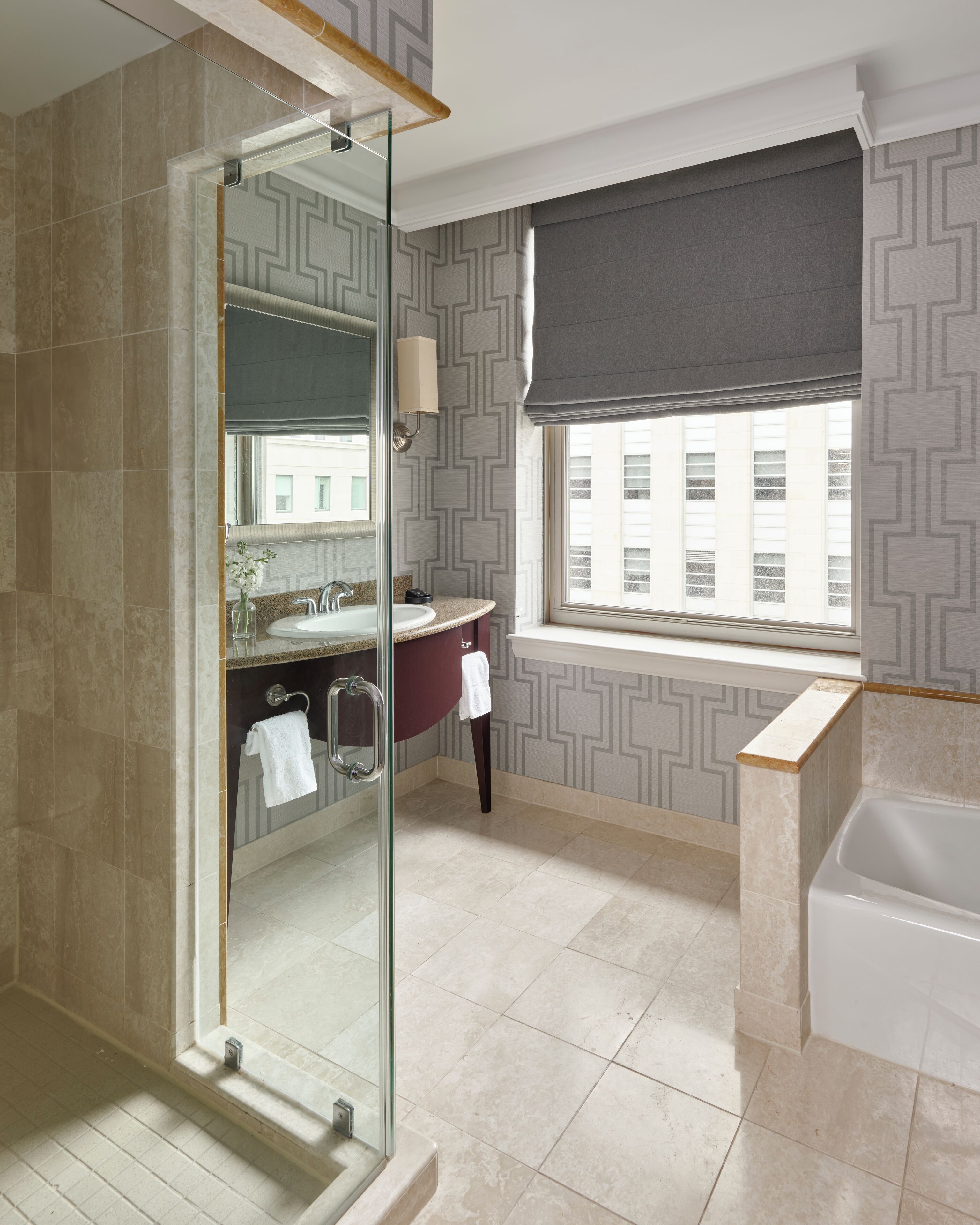 Un baño moderno con cabina de ducha de vidrio, tocador con lavabo y espejo, bañera y ventana con vistas a edificios.