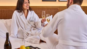 A couple in a hotel room, sitting on a bed with a bottle of wine and a glass of champagne.