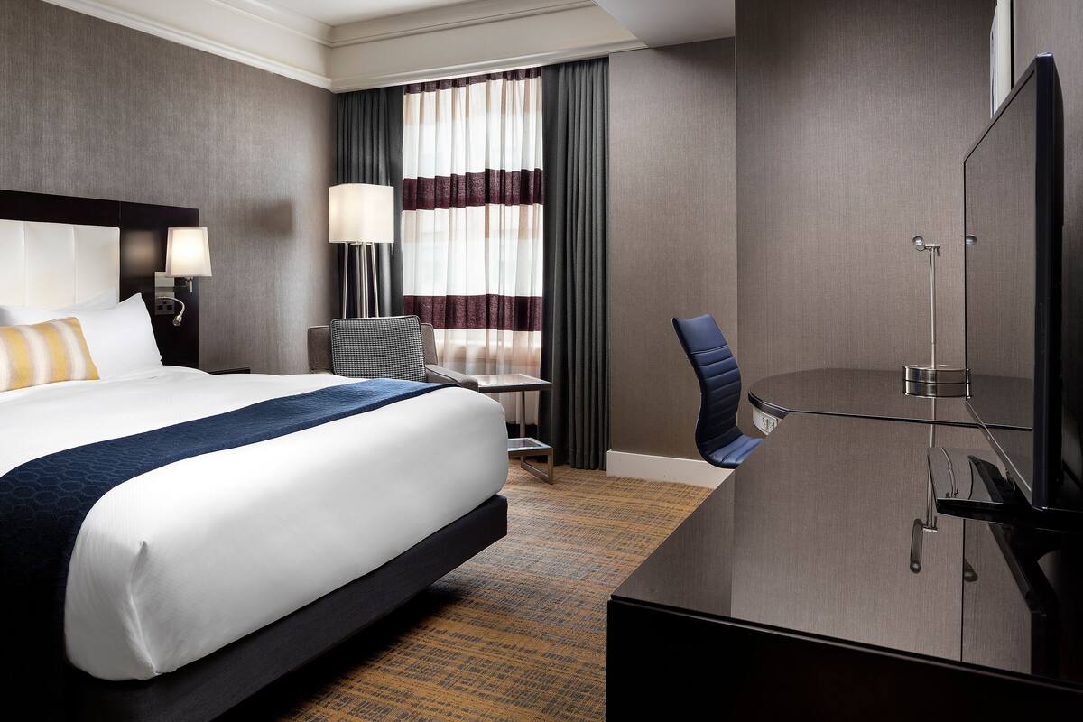 A hotel room with a large bed, a desk, a chair, a TV, and a window with curtains.