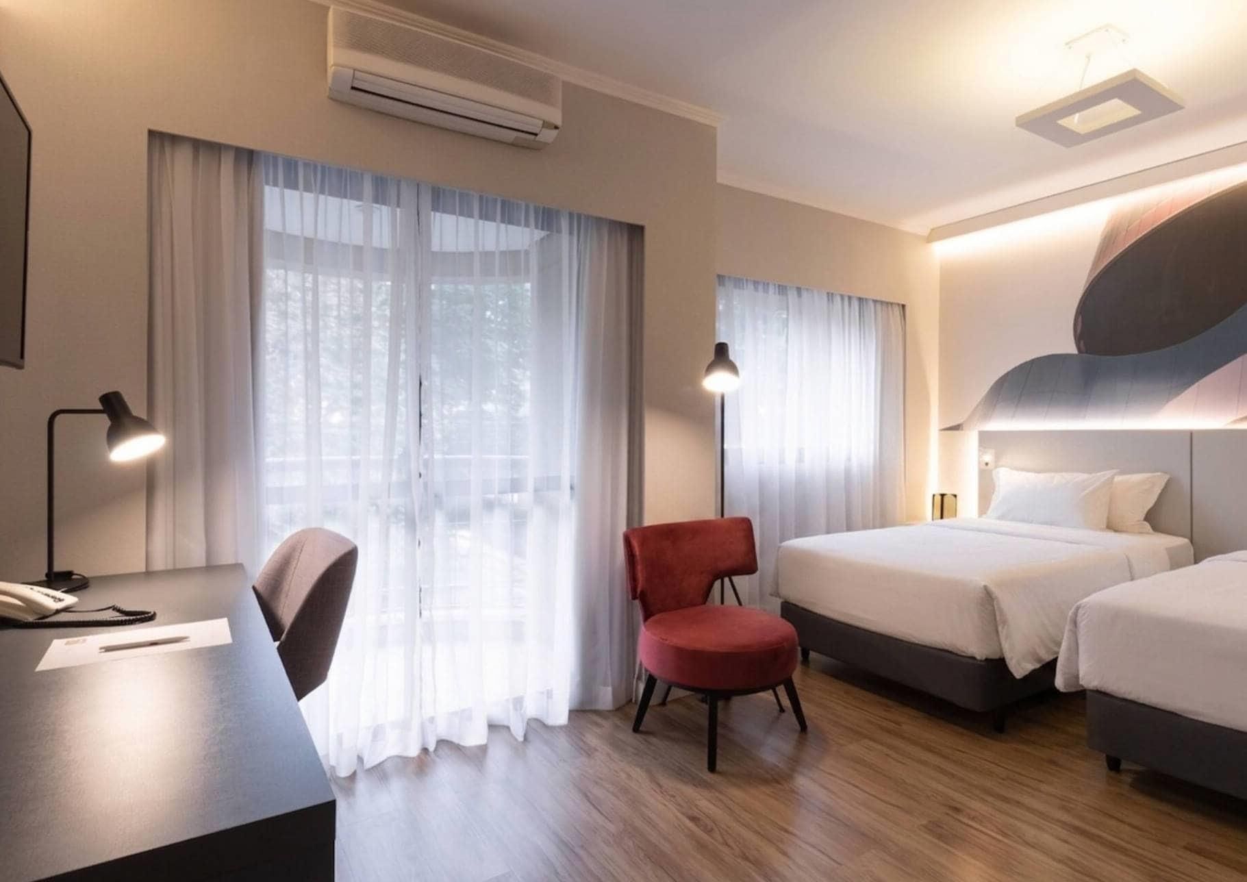 Une chambre d’hôtel équipée d’un bureau, de deux lits, d’une chaise et de grandes fenêtres avec des rideaux légers.