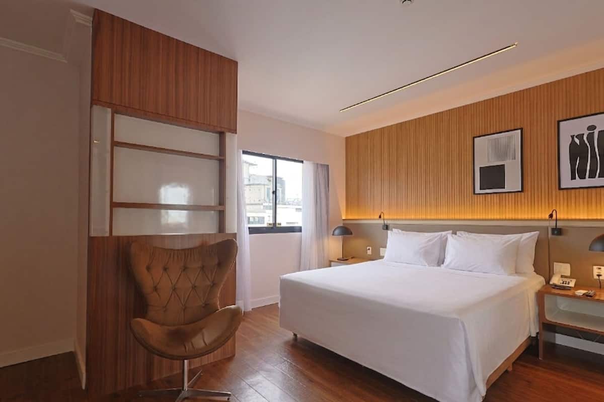 A hotel room with a bed, a chair, a nightstand, a lamp, and a wall-mounted shelf.