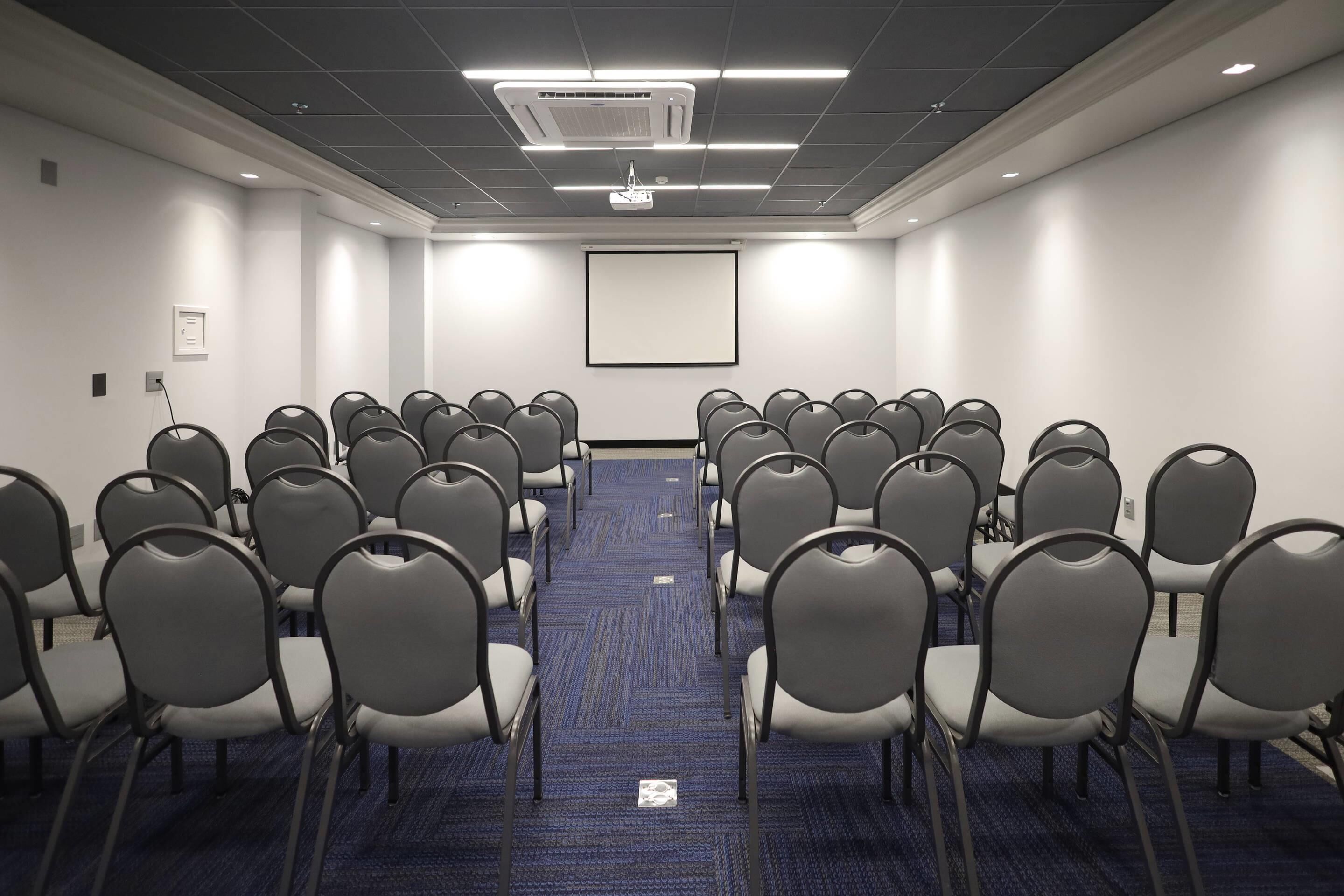 A conference room with rows of chairs, a projector screen, and a carpeted floor.