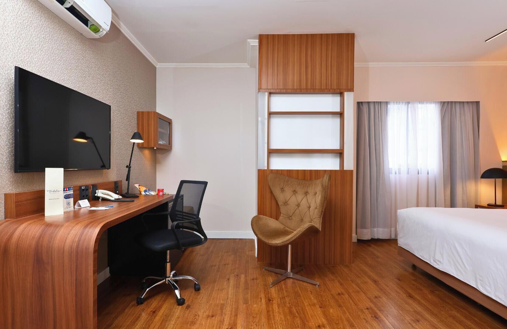Una habitación de hotel con un escritorio de madera, una silla, una cama, un televisor y una ventana con cortinas.