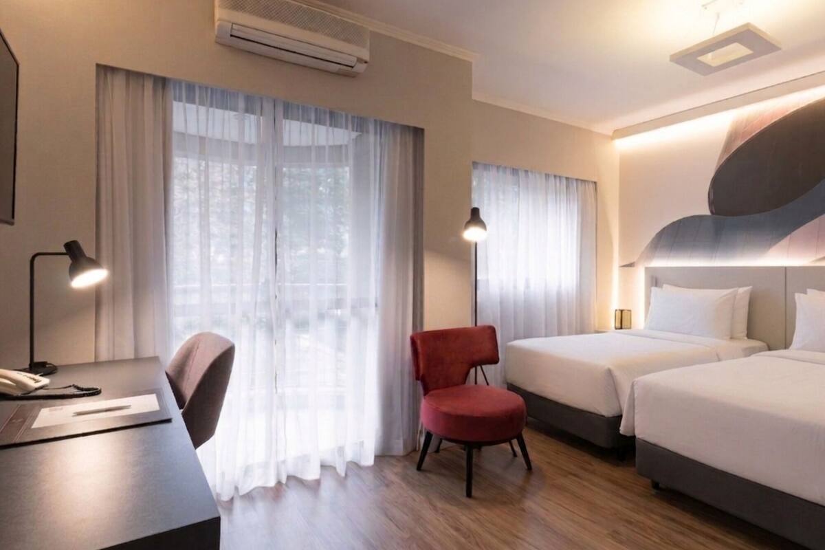 A hotel room with a desk, two beds, a red chair, and a large window with curtains.