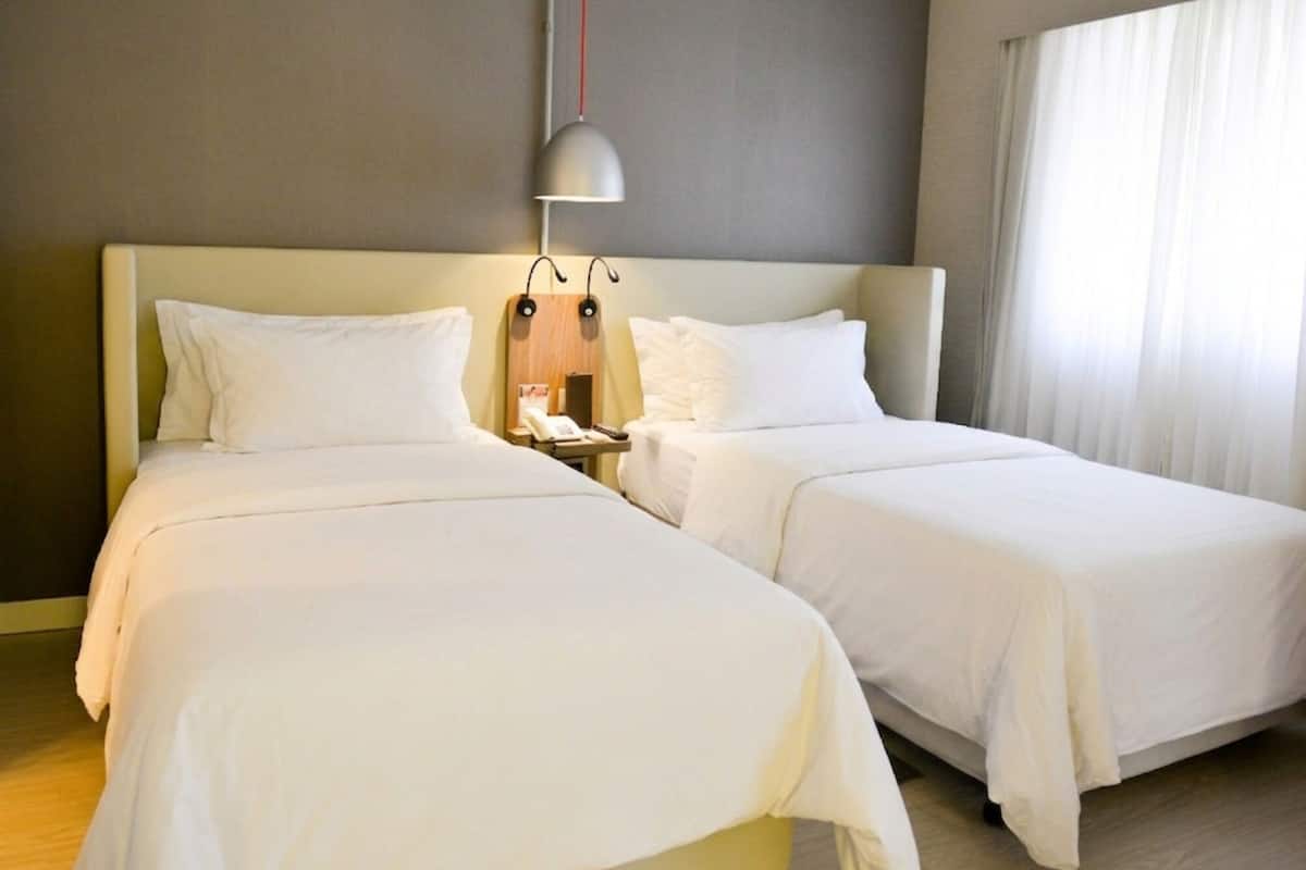A hotel room with two beds, a bedside table with a lamp, and a window with curtains.