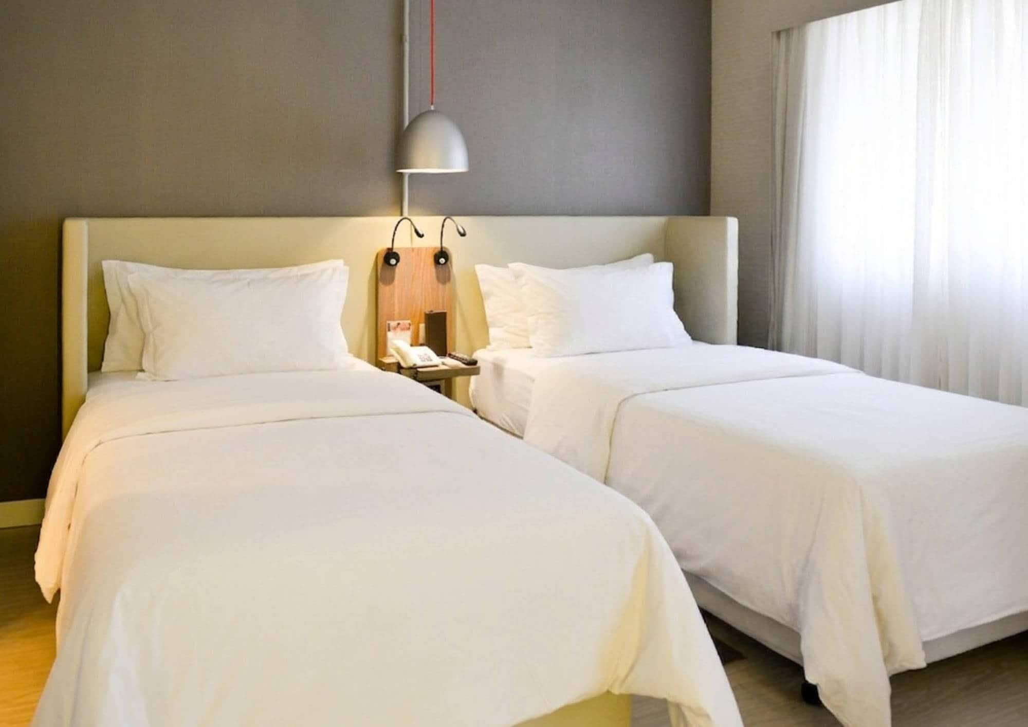 A hotel room with two single beds, a bedside table with a lamp, and a window with curtains.