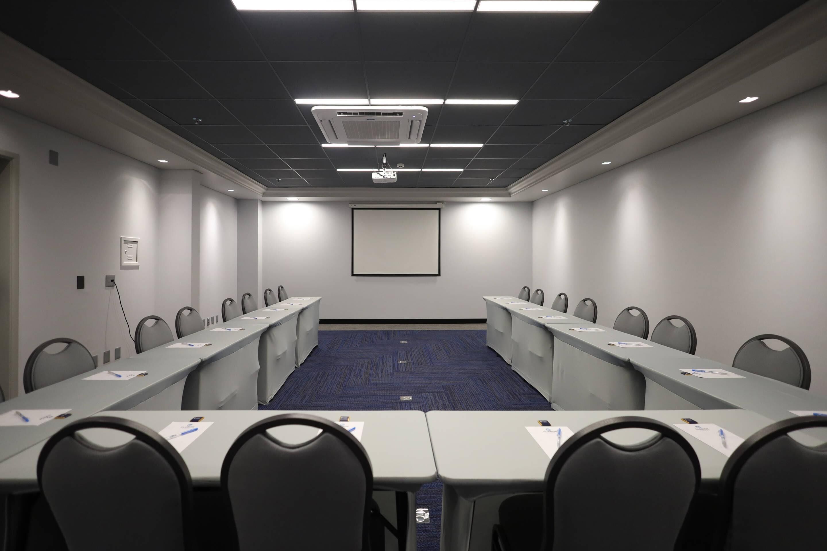 A conference room with a long table, chairs, a projector screen, and a carpeted floor.