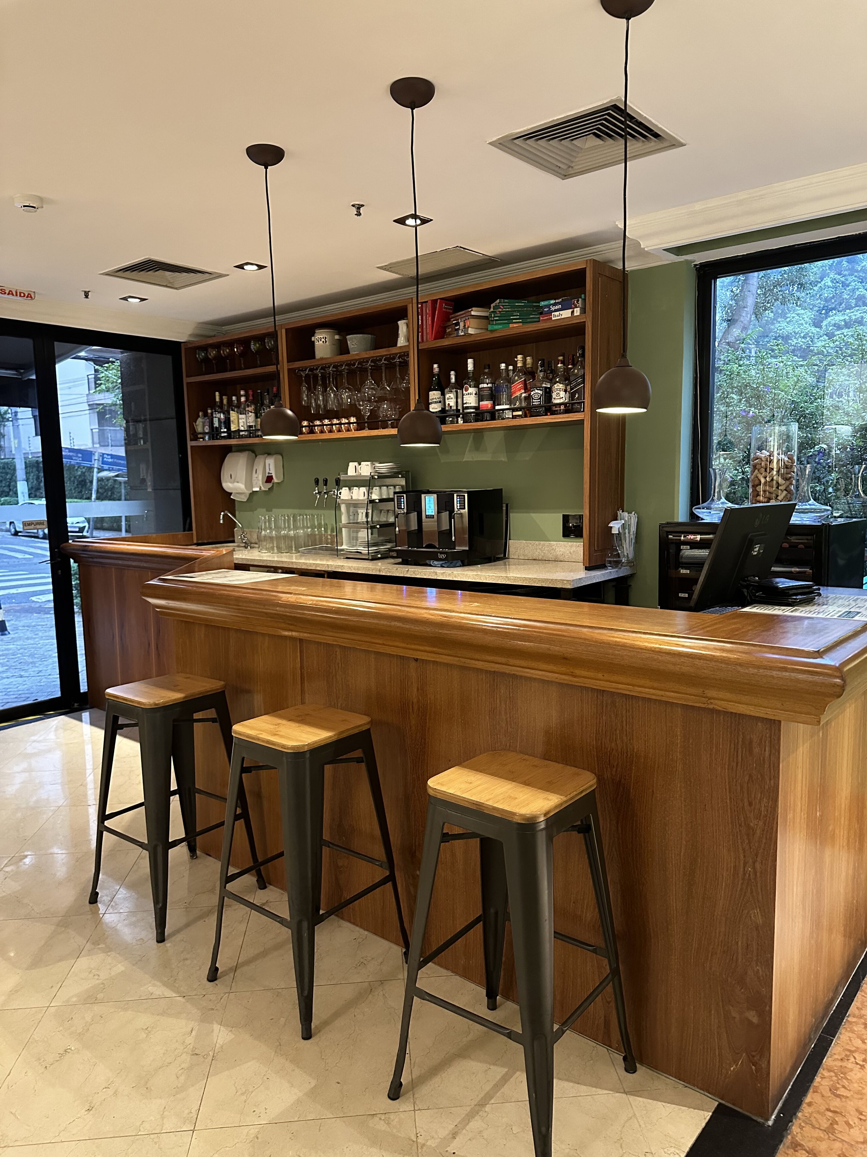 A bar area with a wooden counter, three stools, a coffee machine, and shelves with bottles and glasses.