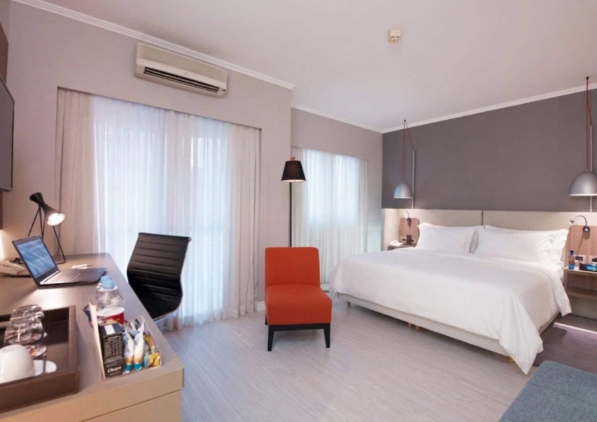 A hotel room with a bed, a desk with a laptop, a chair, a bedside table, and a lamp.
