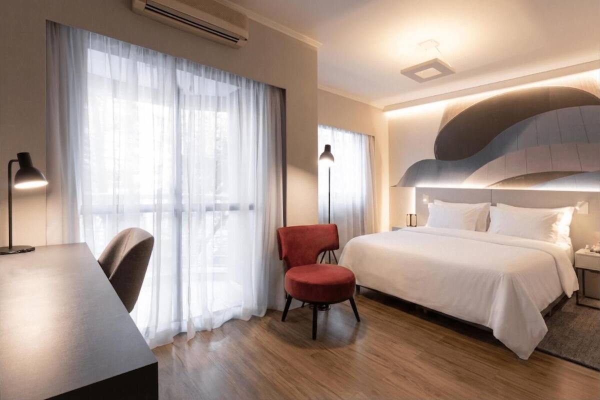 A hotel room with a bed, a desk, a chair, and a large window with sheer curtains.