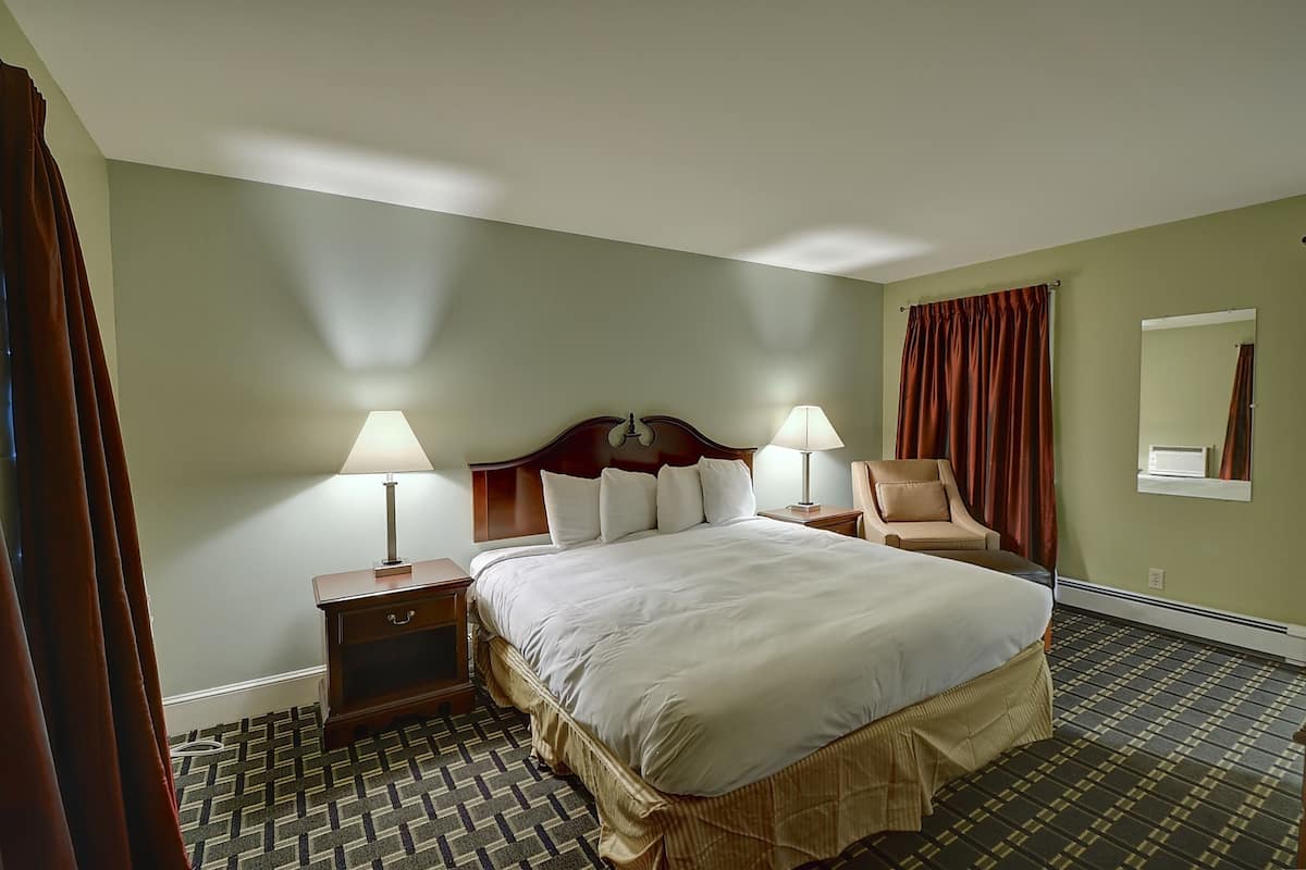 Quarto de hotel com uma cama grande, duas mesas de cabeceira com abajures, uma cadeira, um espelho e um tapete estampado.
