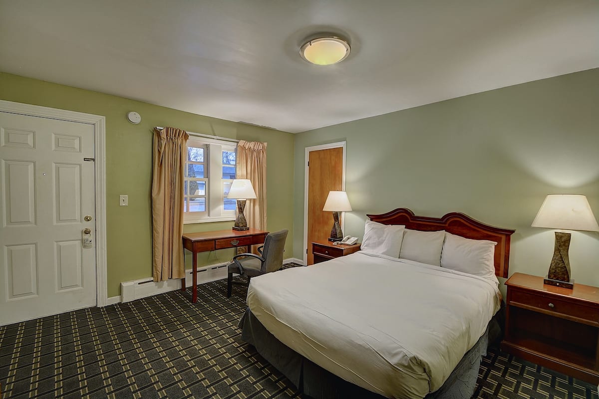 Quarto de hotel com uma cama, duas mesas de cabeceira com abajures, uma escrivaninha com cadeira, uma janela com cortinas e uma porta.