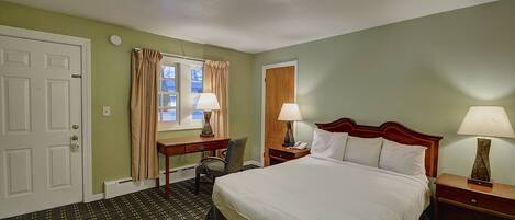 A hotel room with a bed, two bedside tables with lamps, a desk with a chair, a window with curtains, and a door.