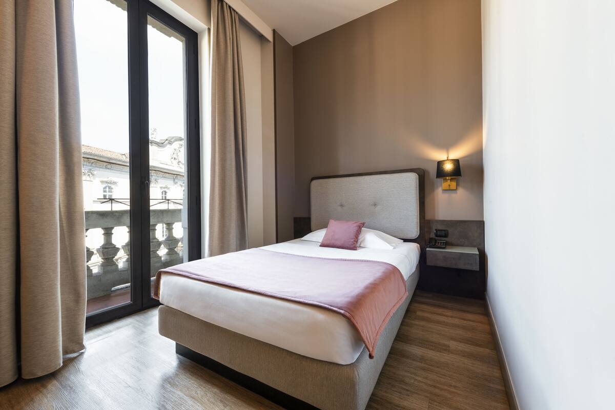 A modern hotel room with a large bed, a nightstand, a wall-mounted lamp, and a view of a balcony.