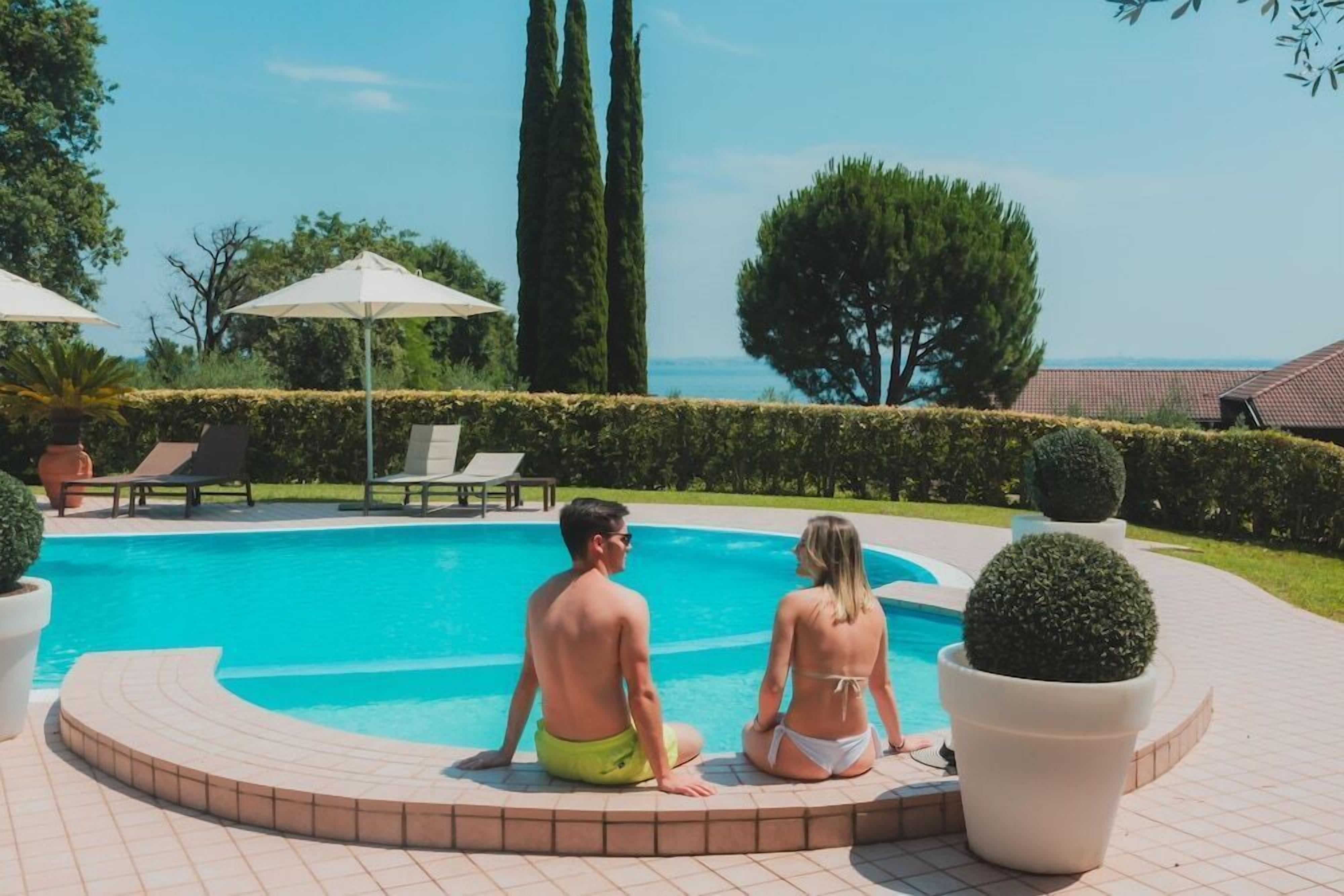 Una parella asseguda a la piscina amb vista al mar, envoltada de plantes en testos i gandules.