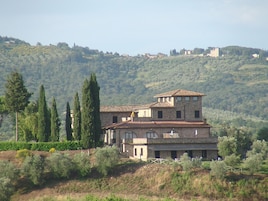 A large, multi-story stone building with a tiled roof, surrounded by trees and hills.