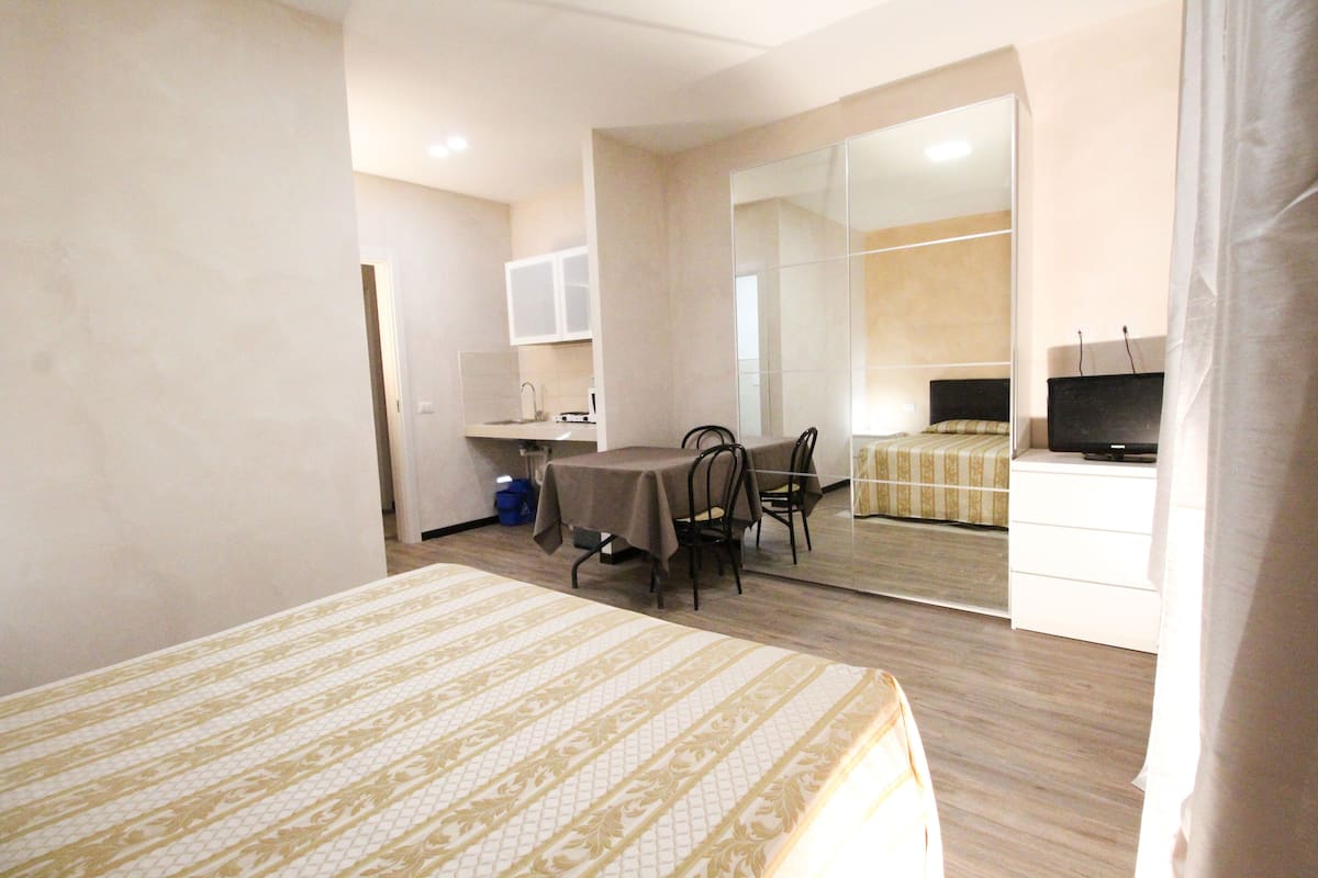 A hotel room with a bed, a dining table with chairs, a small kitchen area, a mirror, and a television.