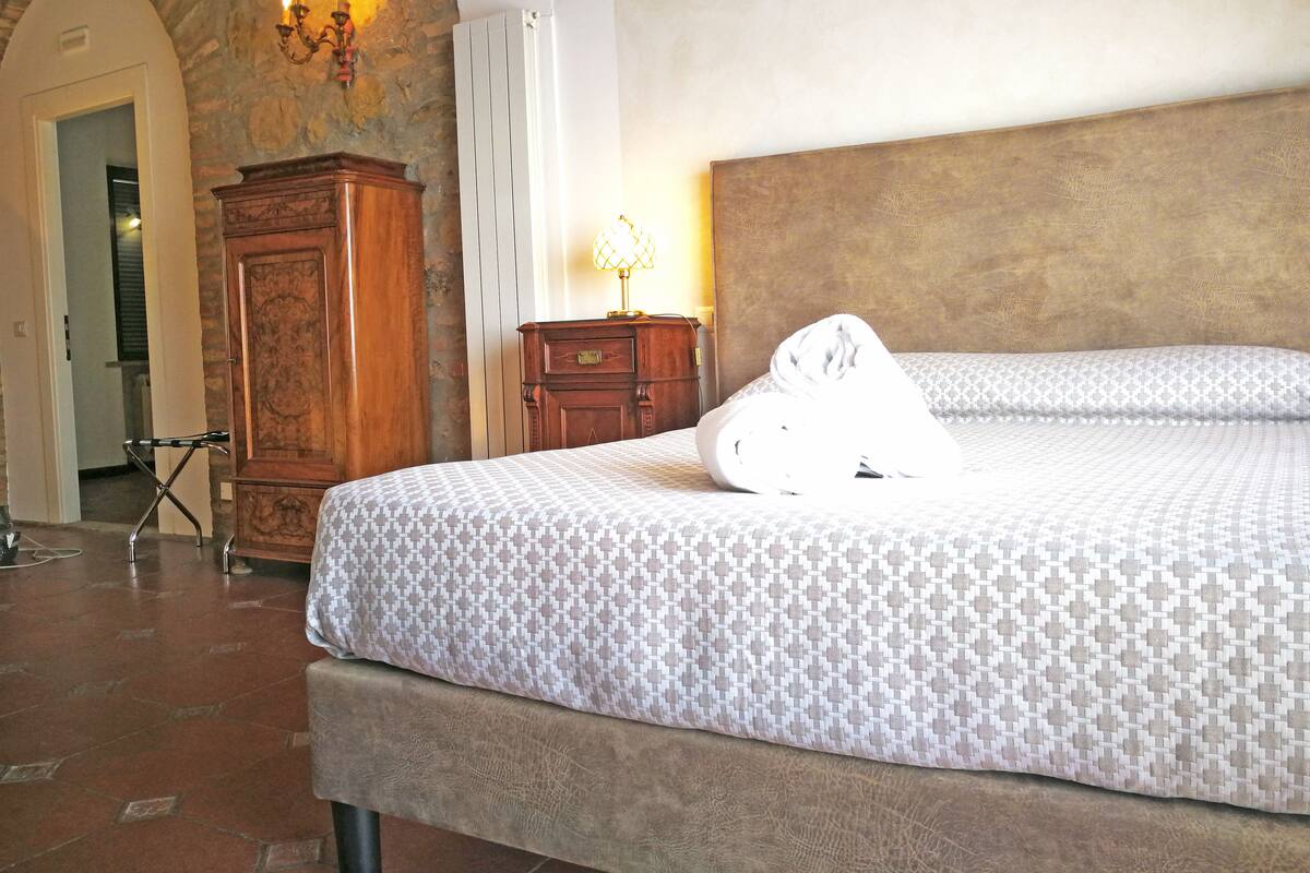 A bedroom with a large bed, a wooden dresser, a stone wall, and a chandelier.