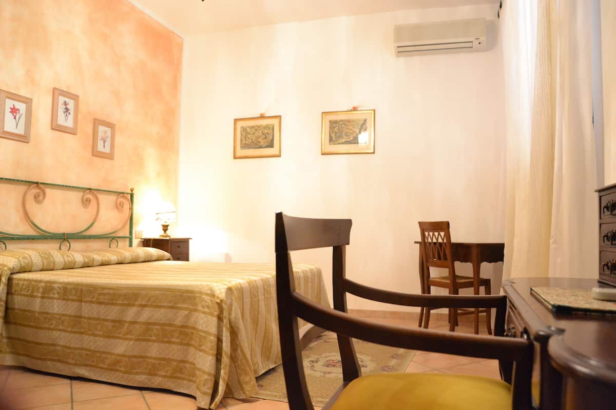 A hotel room with a bed, a chair, a table, and framed pictures on the wall.