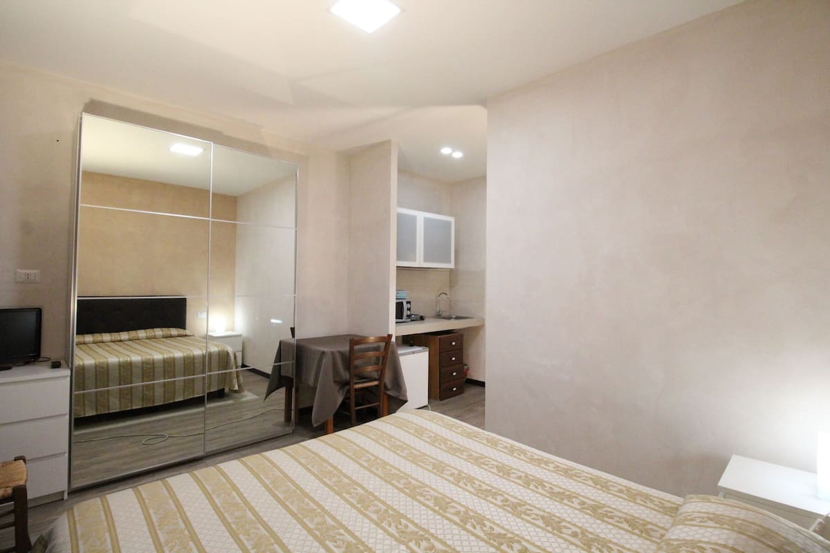 A bedroom with a bed, a desk, a chair, a mirror, a television, and a kitchenette.