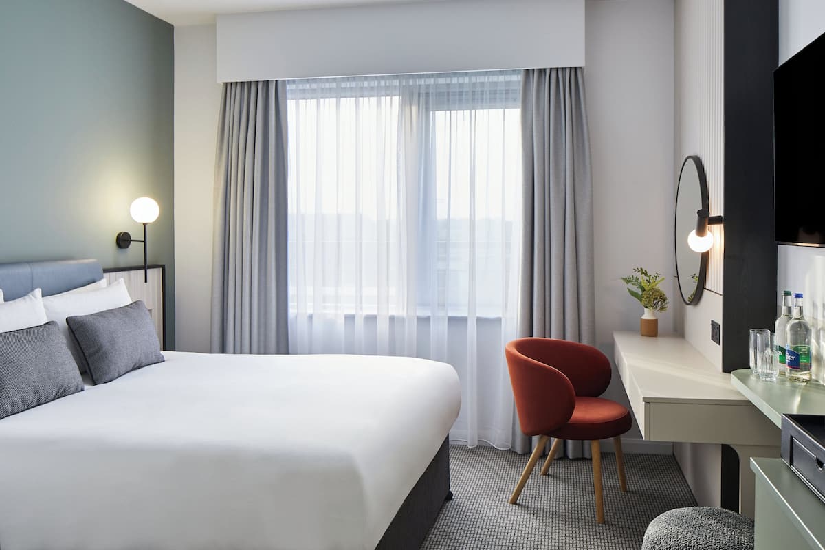 Habitación de hotel moderna con una cama grande, un escritorio, una silla, un espejo y una ventana con cortinas.