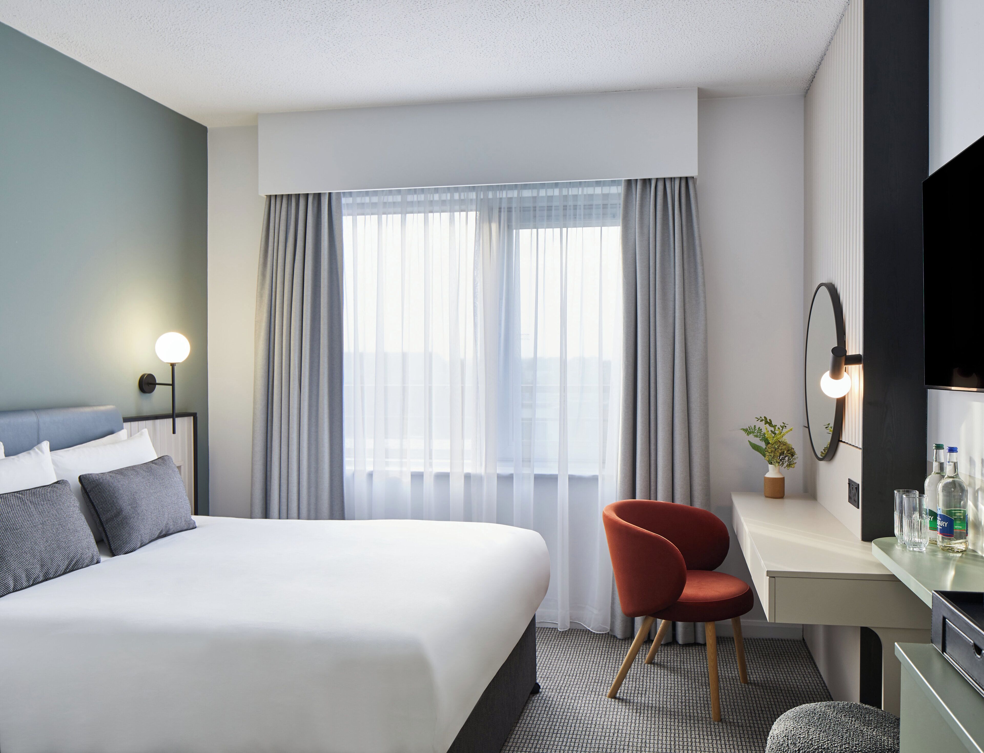 Habitación de hotel moderna con una cama grande, un escritorio, una silla, un espejo y una ventana con cortinas.