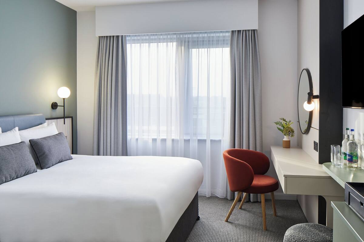 A modern hotel room with a large bed, a desk, a chair, a mirror, and a window with curtains.