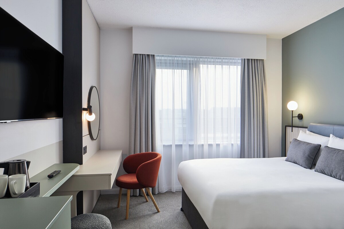A hotel room with a bed, a desk with a chair, a TV, and a window with curtains.