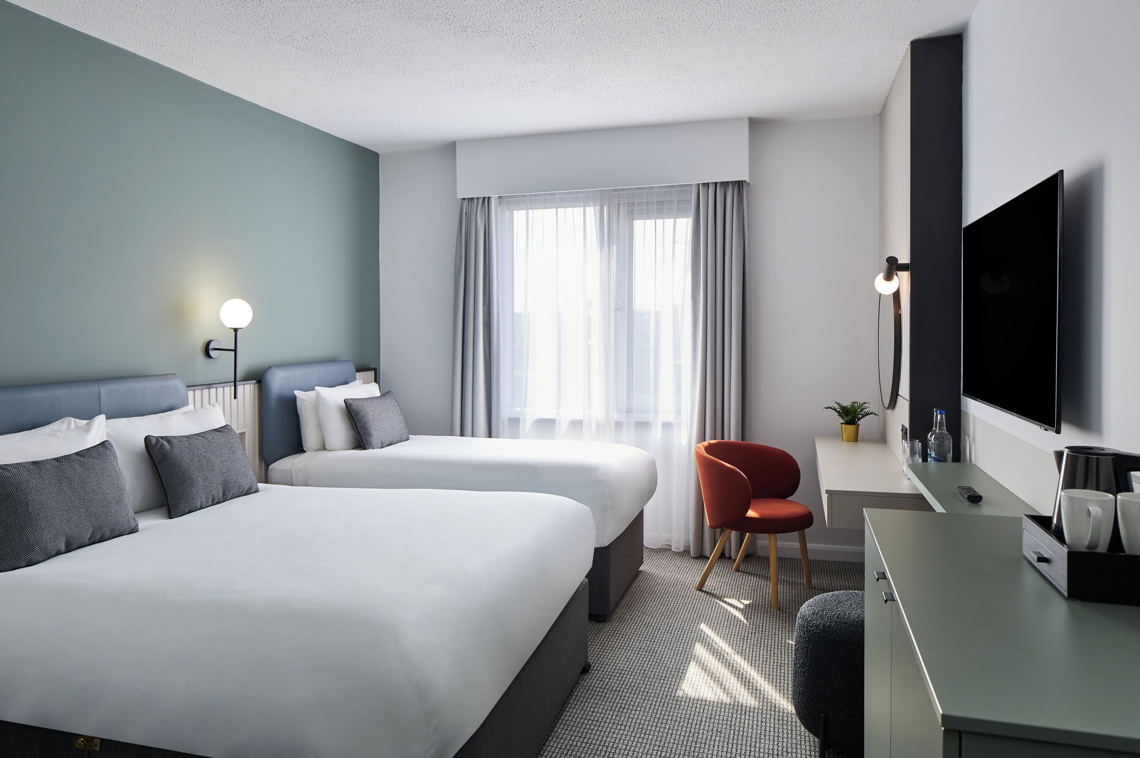 A hotel room with two beds, a desk with a TV, a chair, and a window with curtains.