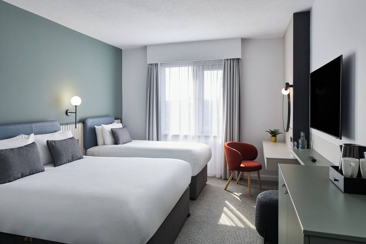 A hotel room with two beds, a desk with a TV, a chair, and a window with curtains.