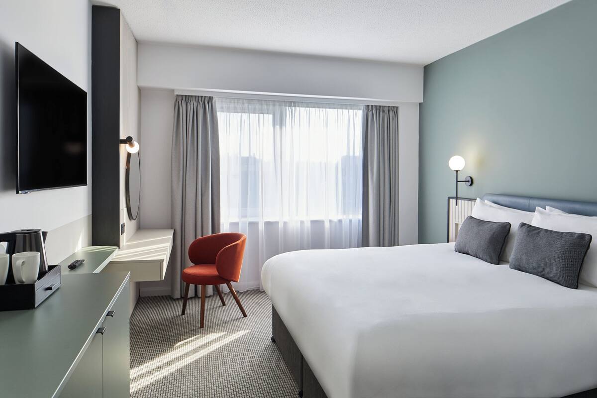 A hotel room with a bed, a desk with a chair, a TV, and a window with curtains.