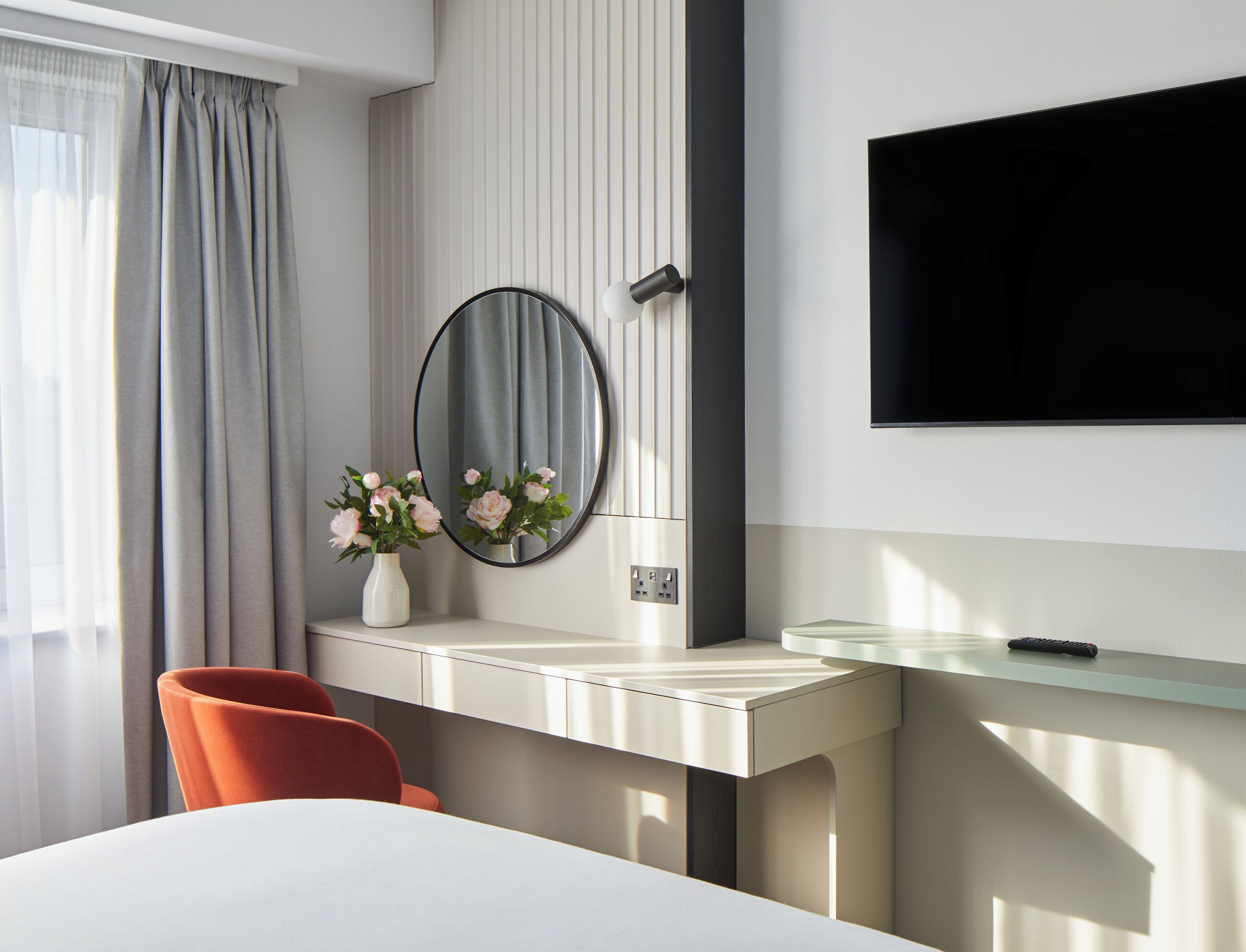 Una habitación de hotel moderna con un escritorio blanco, un espejo redondo, un florero con flores, una silla roja y un televisor montado en la pared.