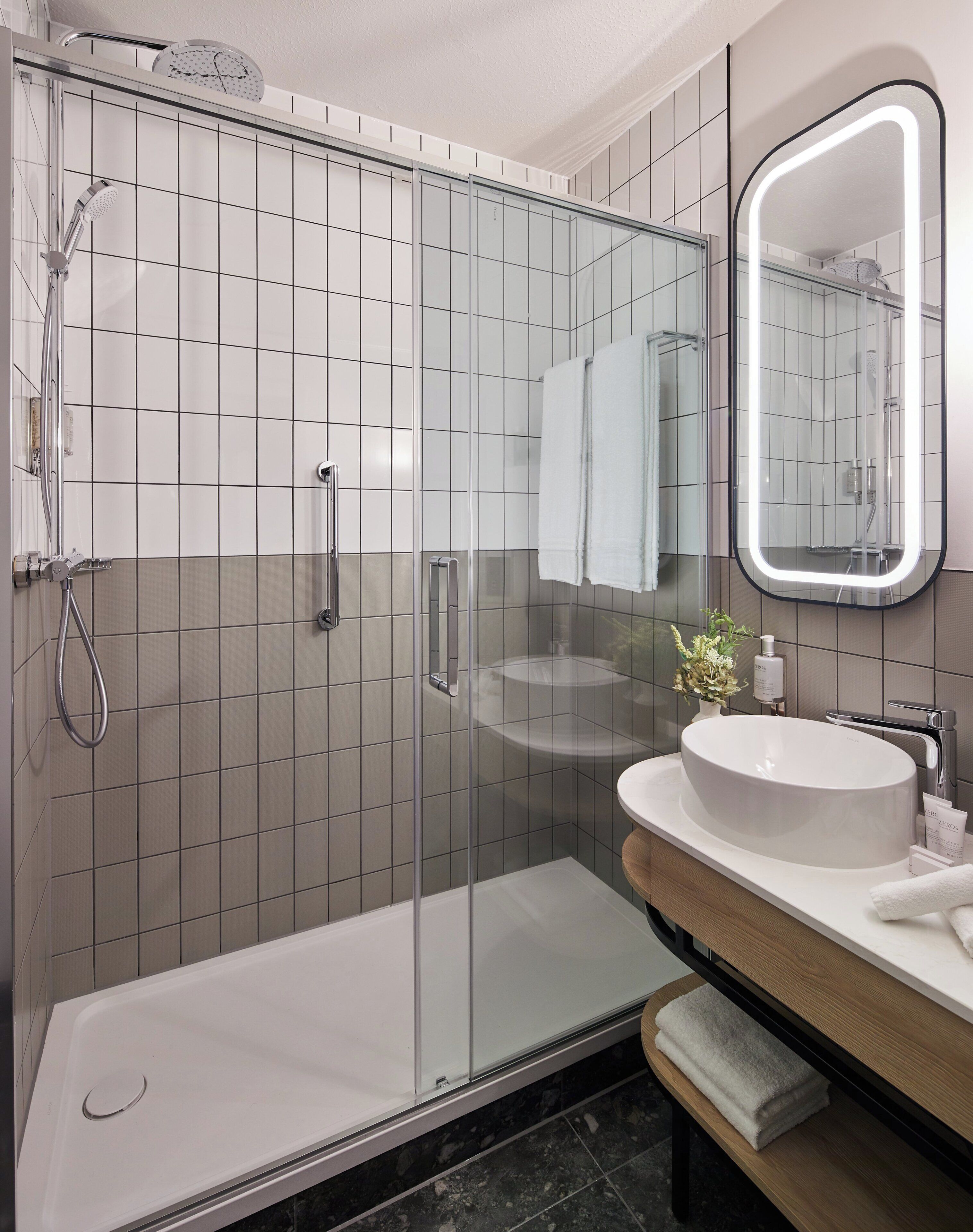 Un baño moderno con ducha de mampara de vidrio, lavamanos blanco y un espejo grande.