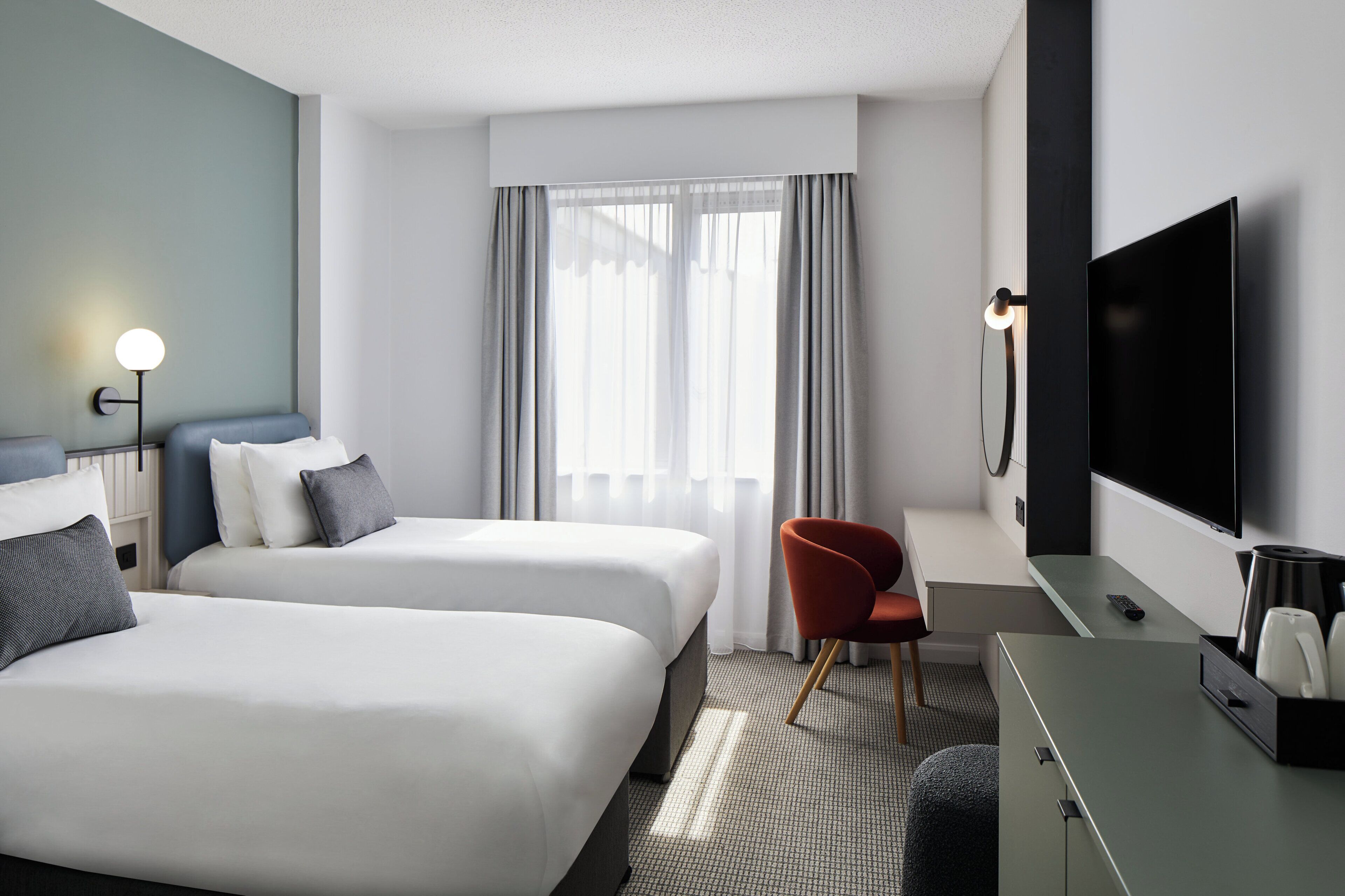 A hotel room with two beds, a desk, a chair, a TV, and a window with curtains.