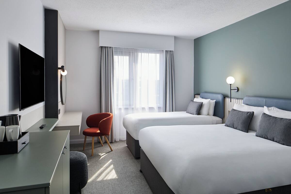 A hotel room with a bed, a desk with a TV, a chair, and a window with curtains.