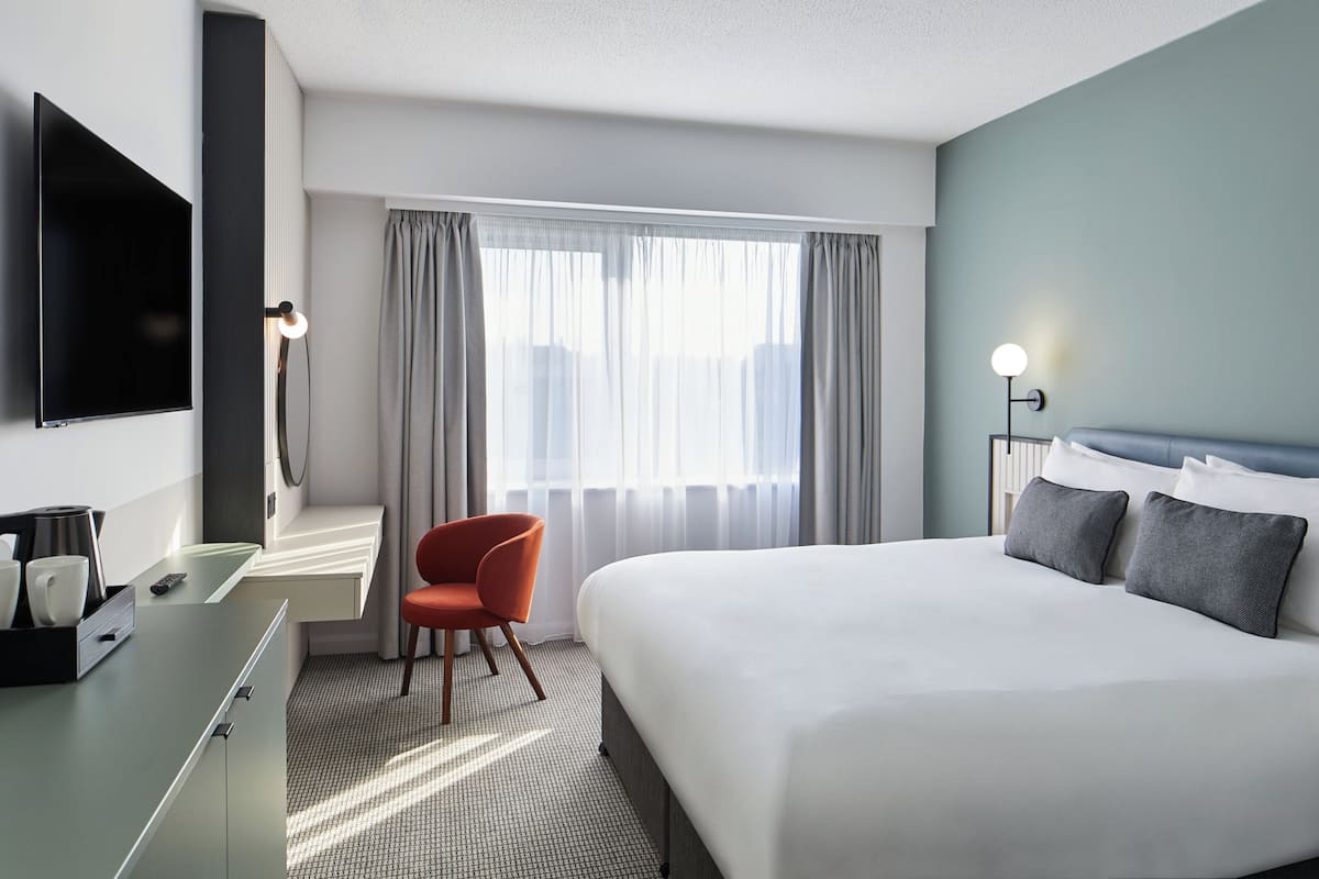 Una habitación de hotel con una cama, un escritorio con silla, un televisor y un ventanal con cortinas.