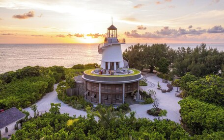 A lighthouse with a circular viewing platform, surrounded by lush greenery and a sandy beach.